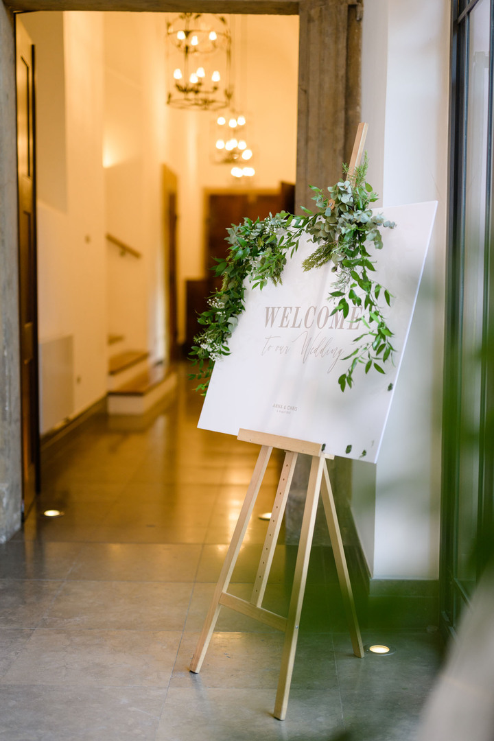 Skleněná tabule "welcome" zdobená zelenou květinou, postavená u vchodu do hotelu Augustine v Praze.