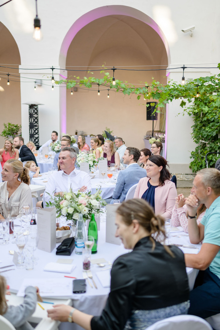 Svatební hosté na venkovní recepci poslouchají proslov a smějí se.