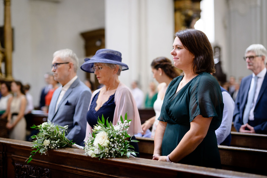 Rodina nevěsty stojící v kostele ve společenských šatech, maminka má modré šaty a modrý klobouk, sestra nevěsty má zelené šaty. Všichni vypadají dojatě.