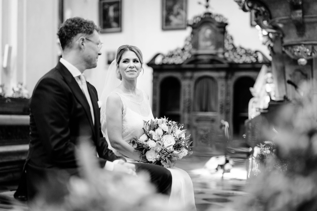 Nevěsta s ženichem sedí na židličkách během obřadu, dívají se na sebe a smějí se. Nevěsta drží na klíně svatební kytici.