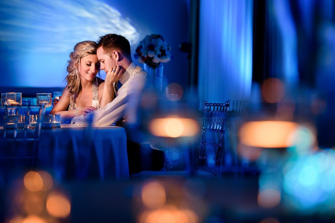 bride and groom sitting at a table with their heads touching in a blue lit room