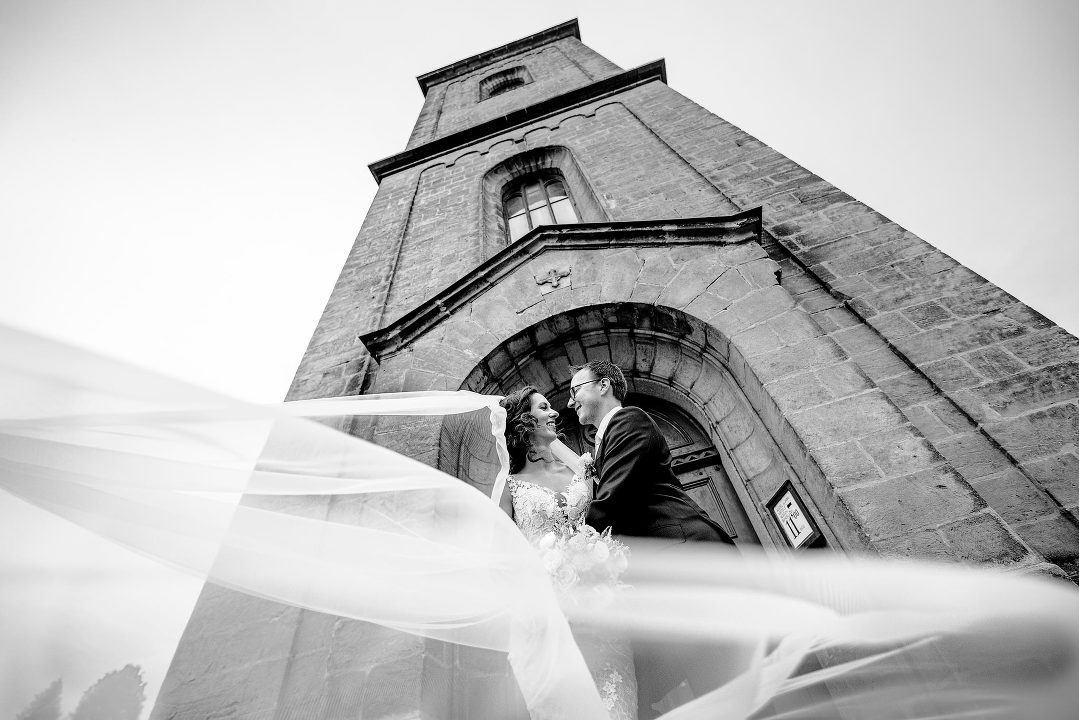 groom holding a bride in white dress with long veil at a church tower