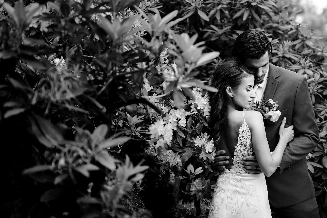 bride in white dress with long hair holding a groom and standing in tall shrub