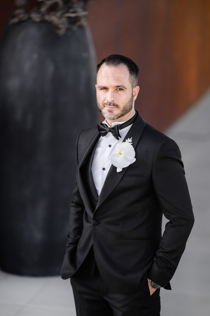 The Groom in black suit with white boutonniere is posing in front of Obelisk Winery.