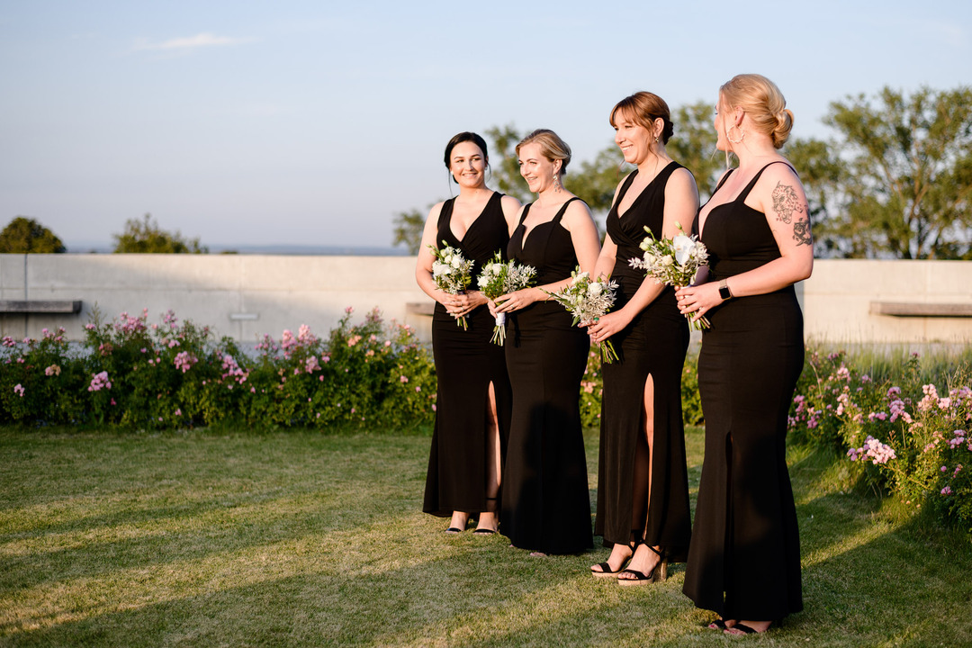 Bridesmaids in elegant black dresses holding bouquets, standing together at an outdoor wedding venue.