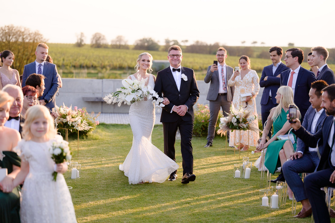 The bride walks down the aisle with her father, while guests watch.