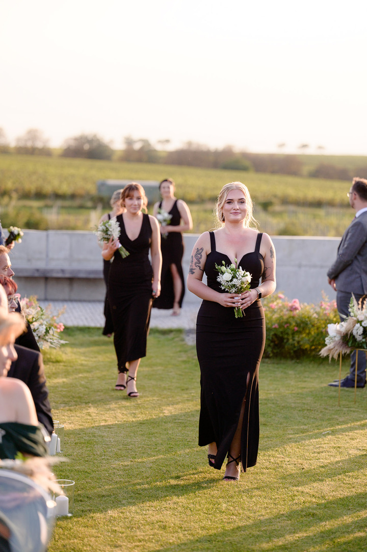 Bridesmaids in elegant black dresses walking down the aisle at Obelisk Winery, holding bouquets.