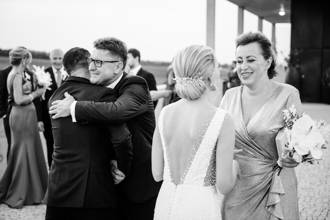 The bride's parents wish the newlyweds well; the mother smiles at the bride emotionally, while the groom's father pats his back and laughs.