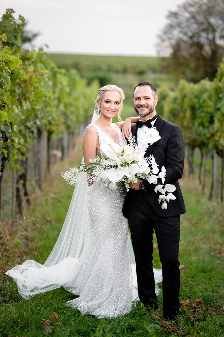 Bride and groom posing together in a vineyard, with the bride holding a bouquet of white flowers.