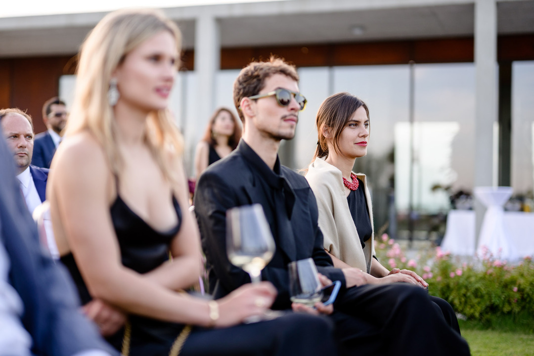 Guests sitting on chairs watch the ceremony, holding a glass of wine.