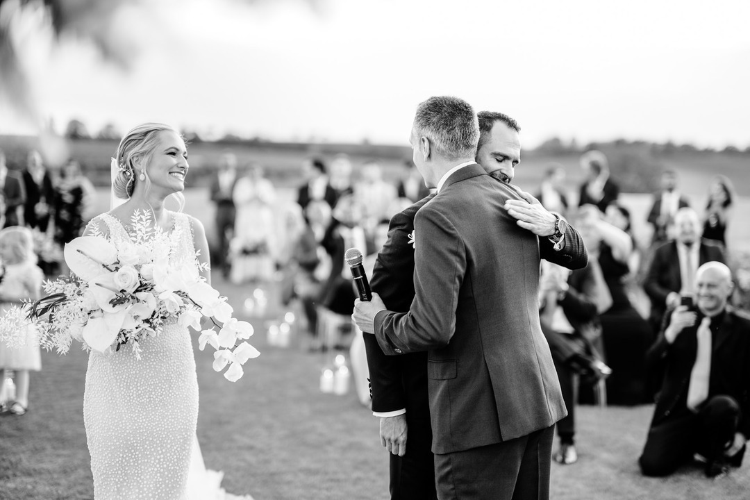 Bride smiles as groom receives a warm hug from his brother and best man during the wedding ceremony at Obelisk Winery.