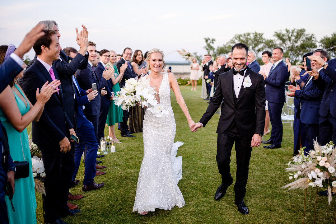 The newlyweds walk down the aisle, surrounded by clapping guests, all smiling.