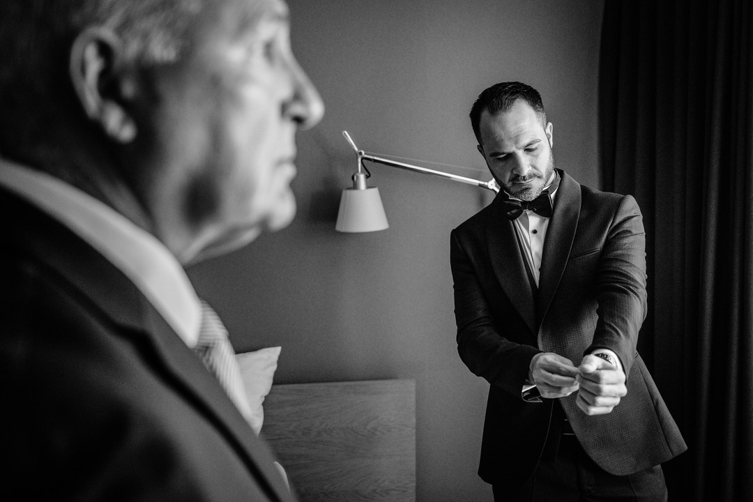 The groom finishes getting ready, fastening his cufflinks. His father stands in the foreground.