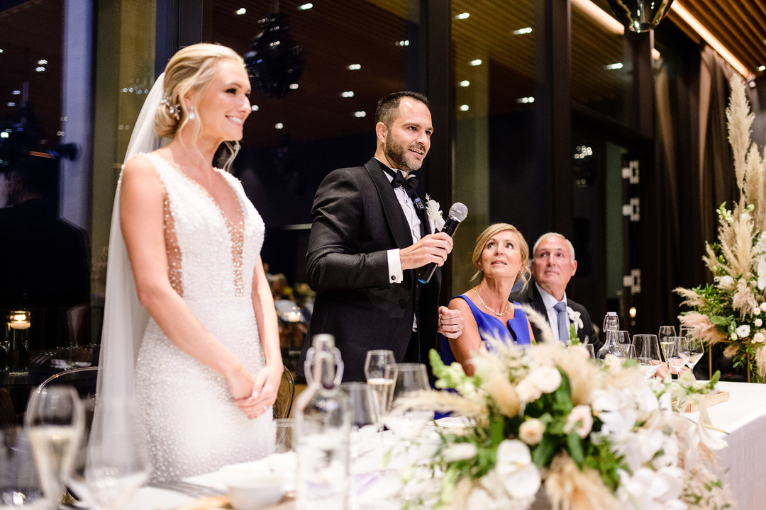 The groom holds a microphone and gives a speech, standing with the bride at the table.