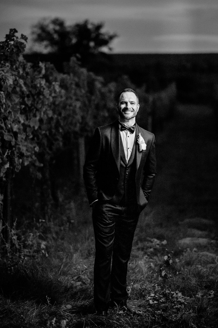 The groom is smiling in vineyard, he has black suit and white boutonniere.