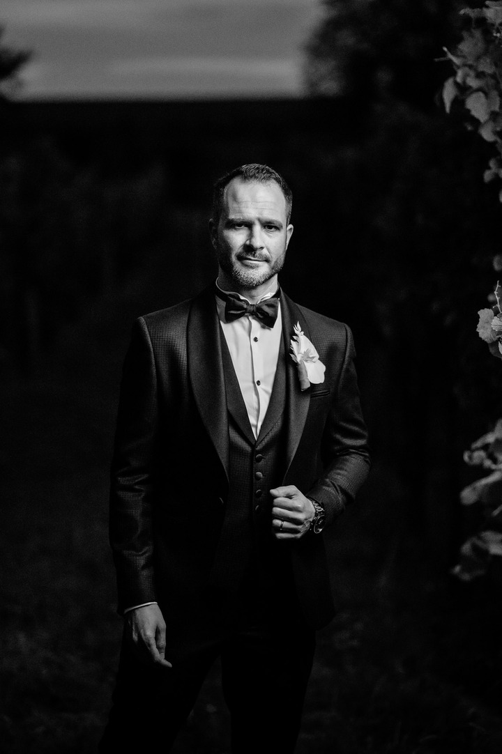 Groom standing confidently in a black tuxedo with a white rose boutonniere at Obelisk Winery.