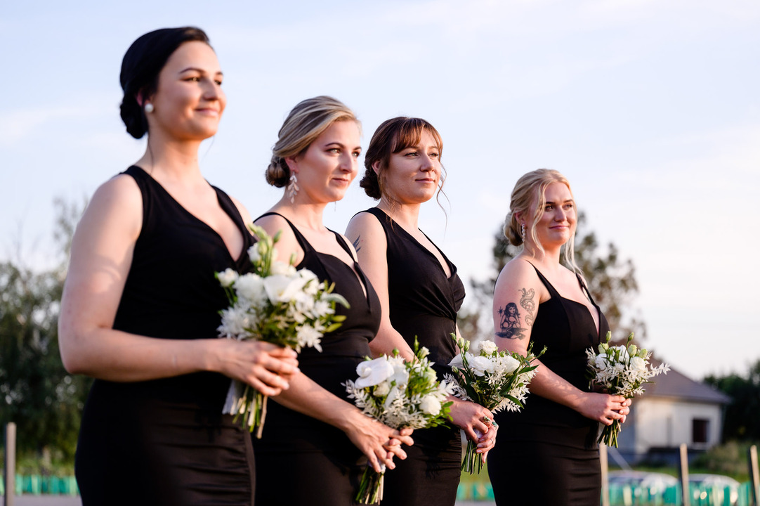 The bride's bridesmaids, all in elegant black dresses, stand side by side, smiling as they watch the ceremony.