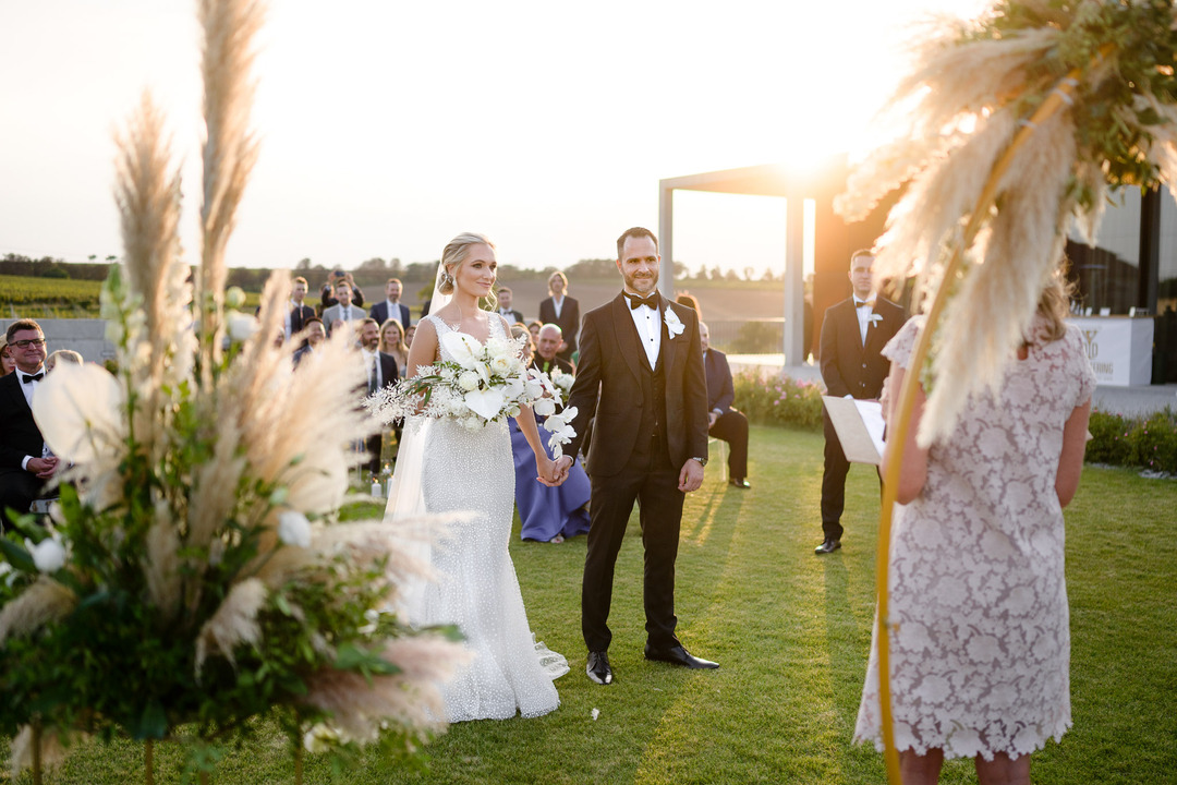 The bride and groom stand hand in hand at the altar, bathed in golden sunlight from behind.