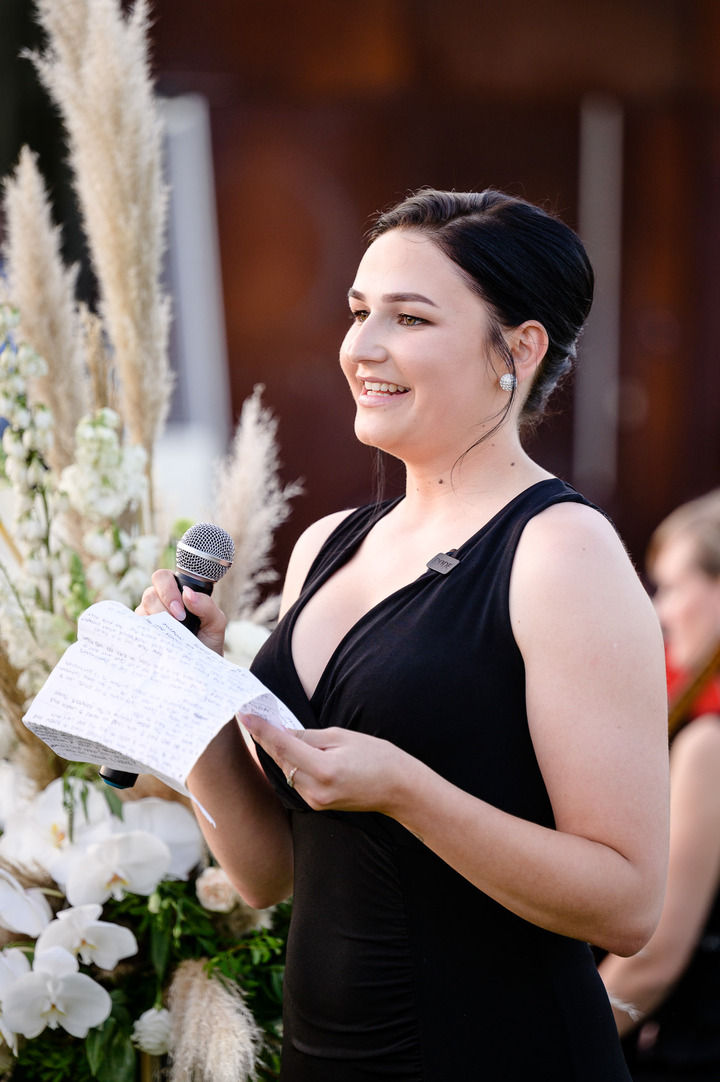 A bridesmaid in a black dress giving a speech at a wedding, holding a microphone.