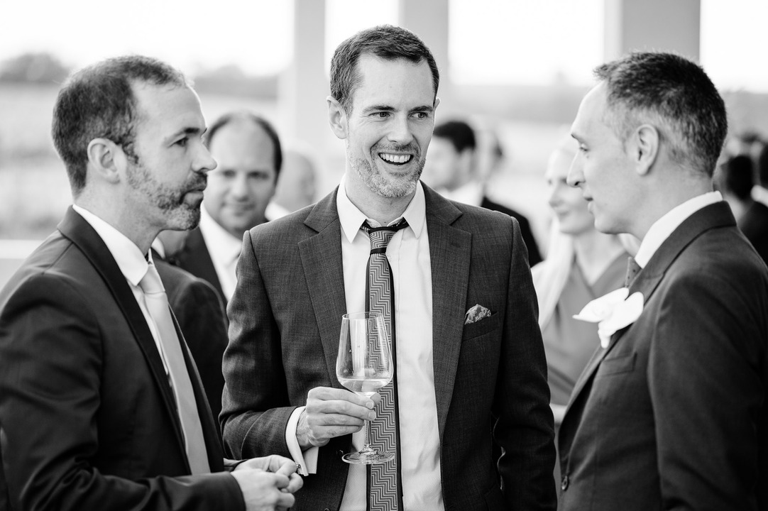 Three men in suits and ties laugh and chat at a wedding reception.