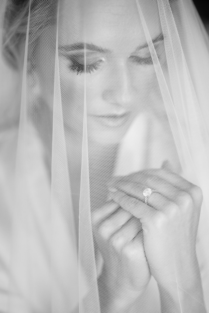 The bride is softly smiling under the veil, she has her engagement ring on her finger. 