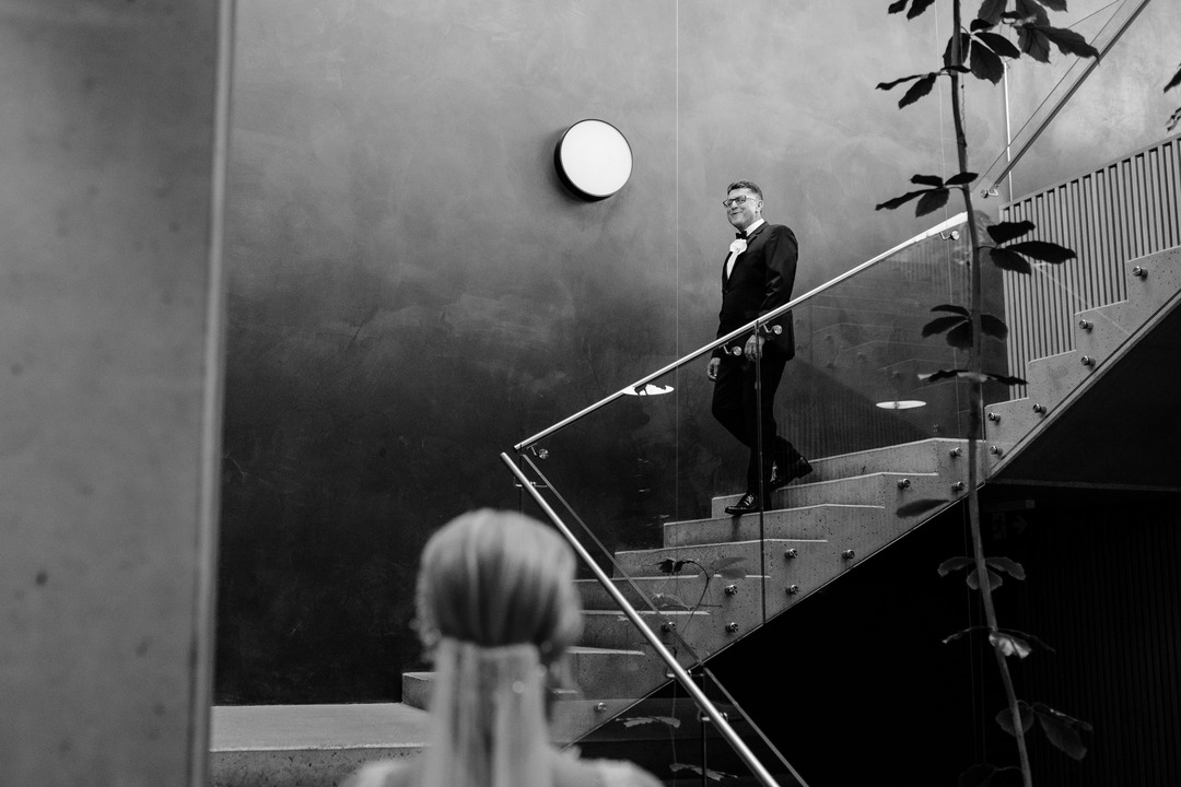 The bride's father standing on a modern staircase, looking at the bride.