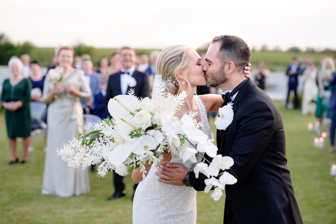 Newlyweds share a kiss at Obelisk Winery, with guests celebrating in the background.