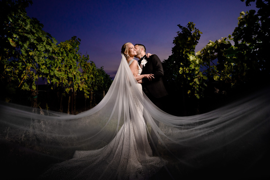 A wide photo of the newlyweds embracing, with the groom kissing the bride on the cheek, and her veil extending into the foreground.