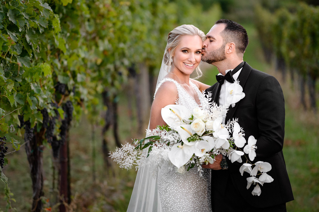 Bride and groom posing together in a vineyard, with the groom kissing the bride at her cheek.