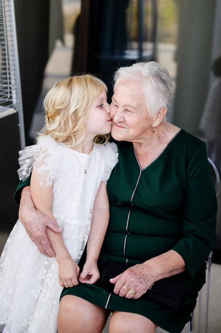 The granddaughter is sitting on her grandmother’s lap and kissing her od the cheek.