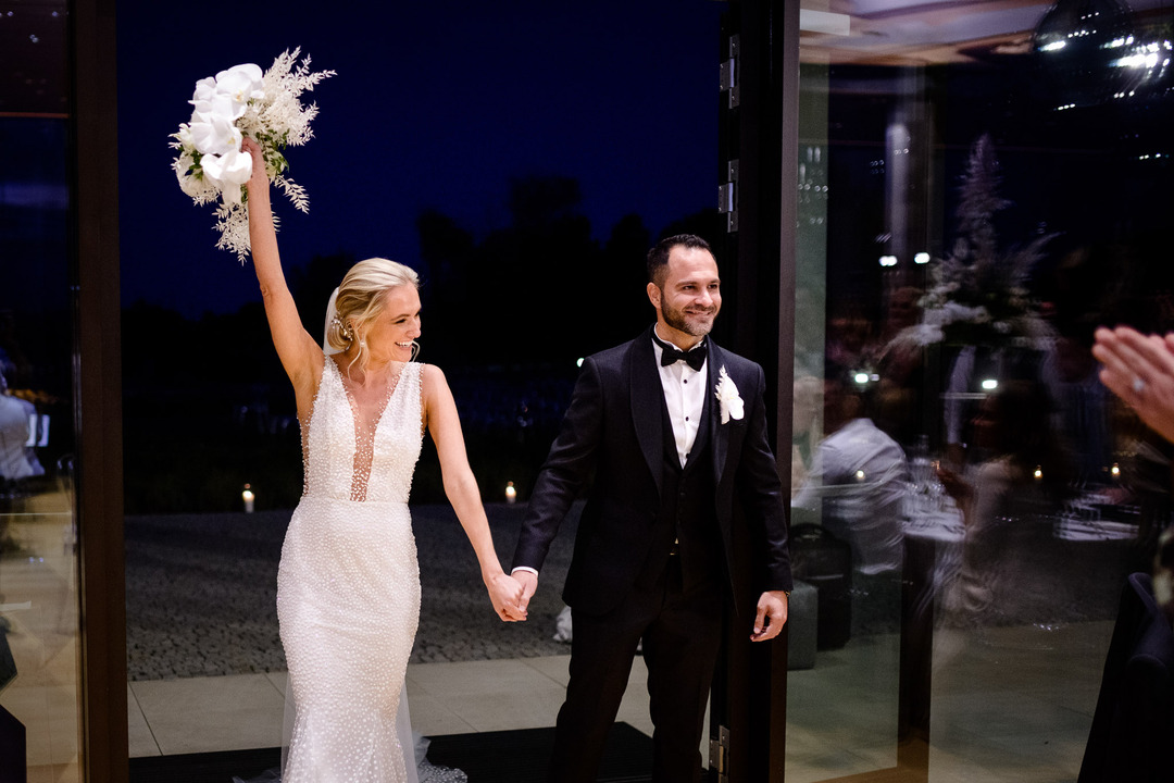 A bride and groom walk hand in hand, waving to guests at their elegant wedding at Obelisk Winery.