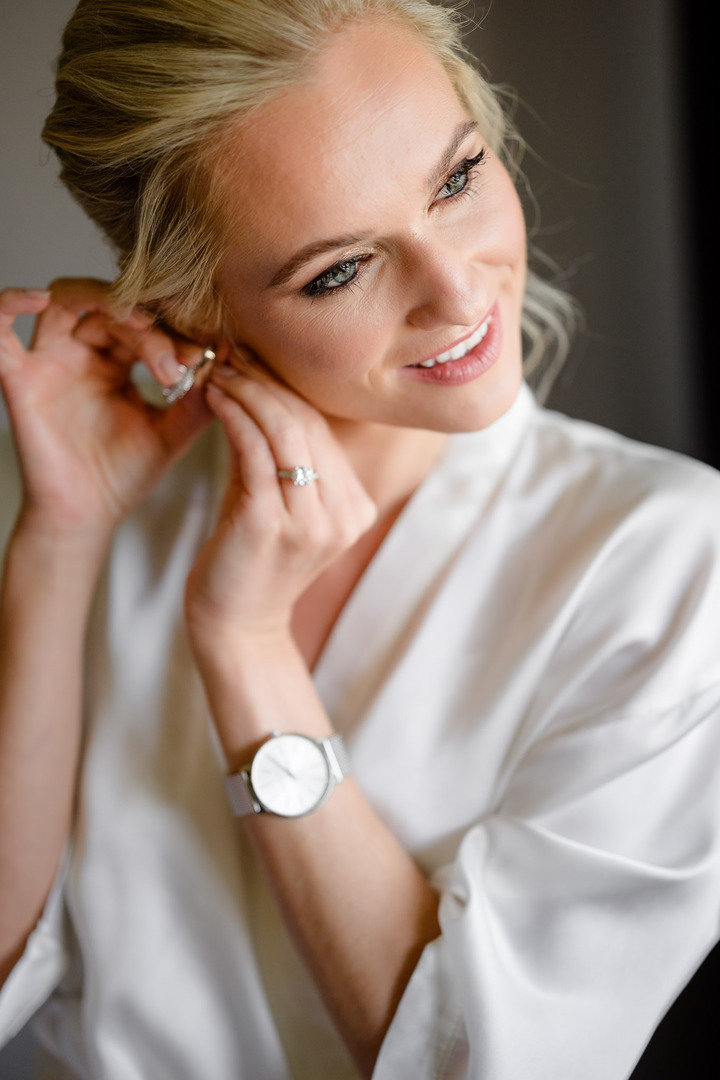 Bride adjusting her earring while wearing a white robe and watch, preparing for her wedding.