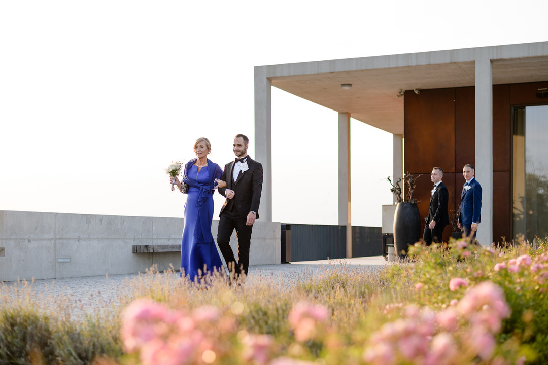 Groom and mother walking arm in arm towards the wedding ceremony at Obelisk Winery, with roses in the foreground.