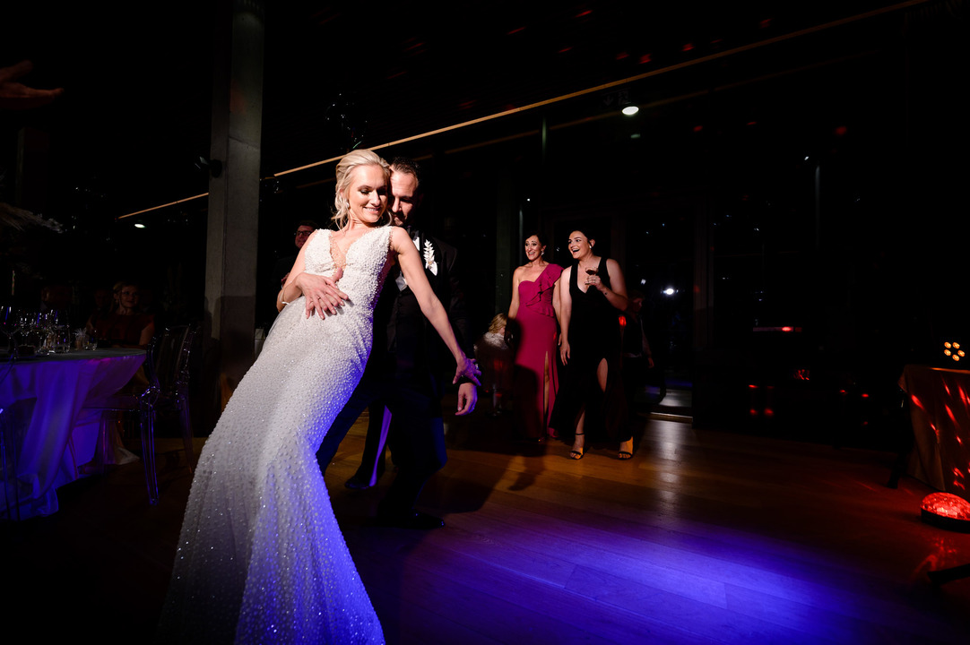 The newlyweds dance on the floor, lit by blue light, with the bride standing with her back to the groom.