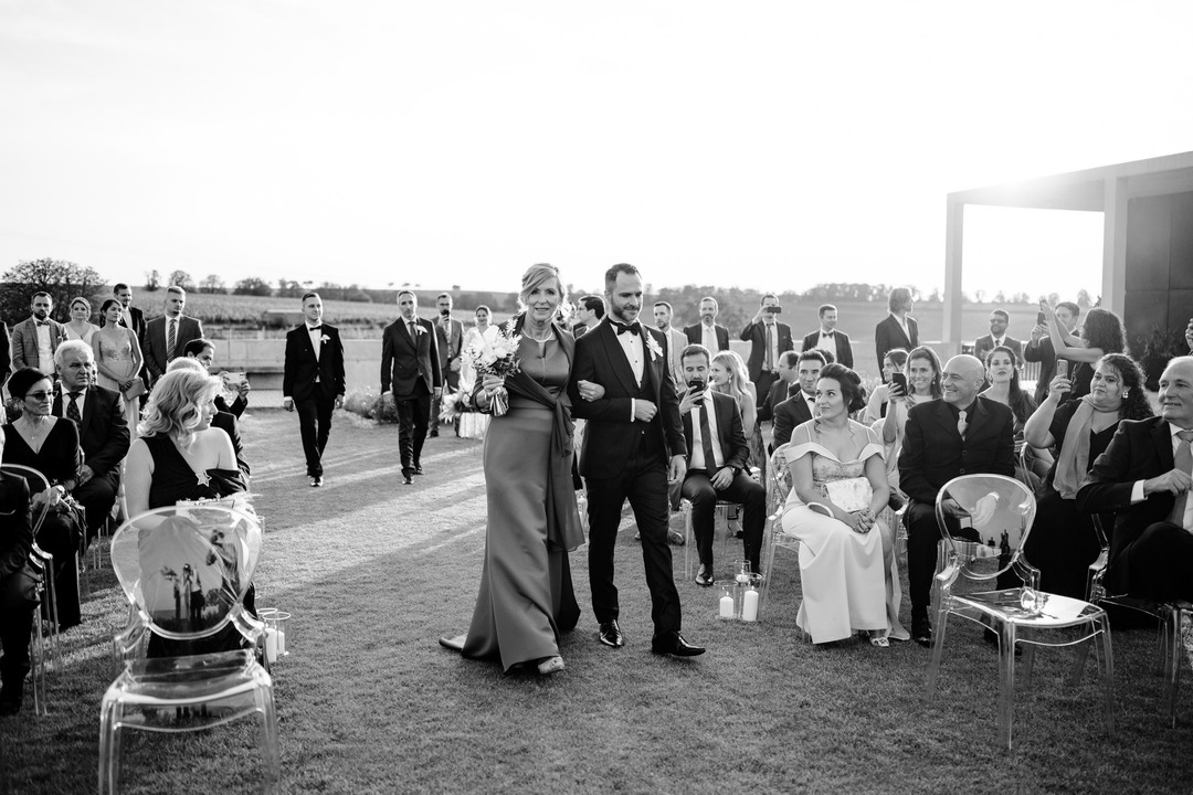 Groom walking with his mother, guests watching and smiling.