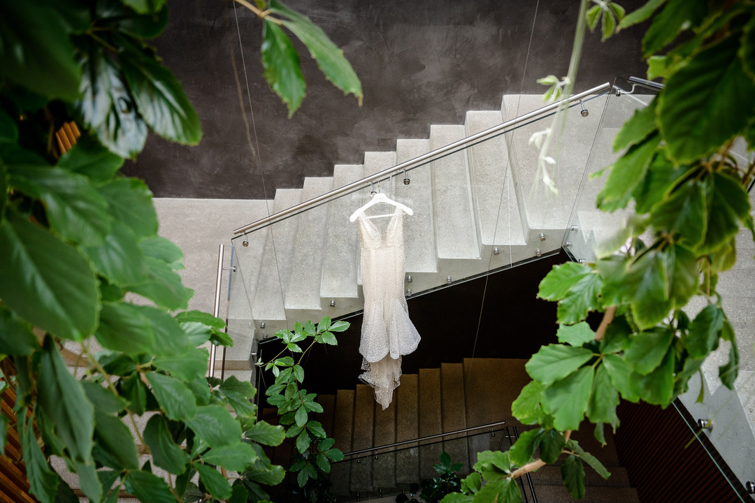 A wedding dress hanging on a glass staircase railing, surrounded by greenery, showcasing intricate lace detailing.