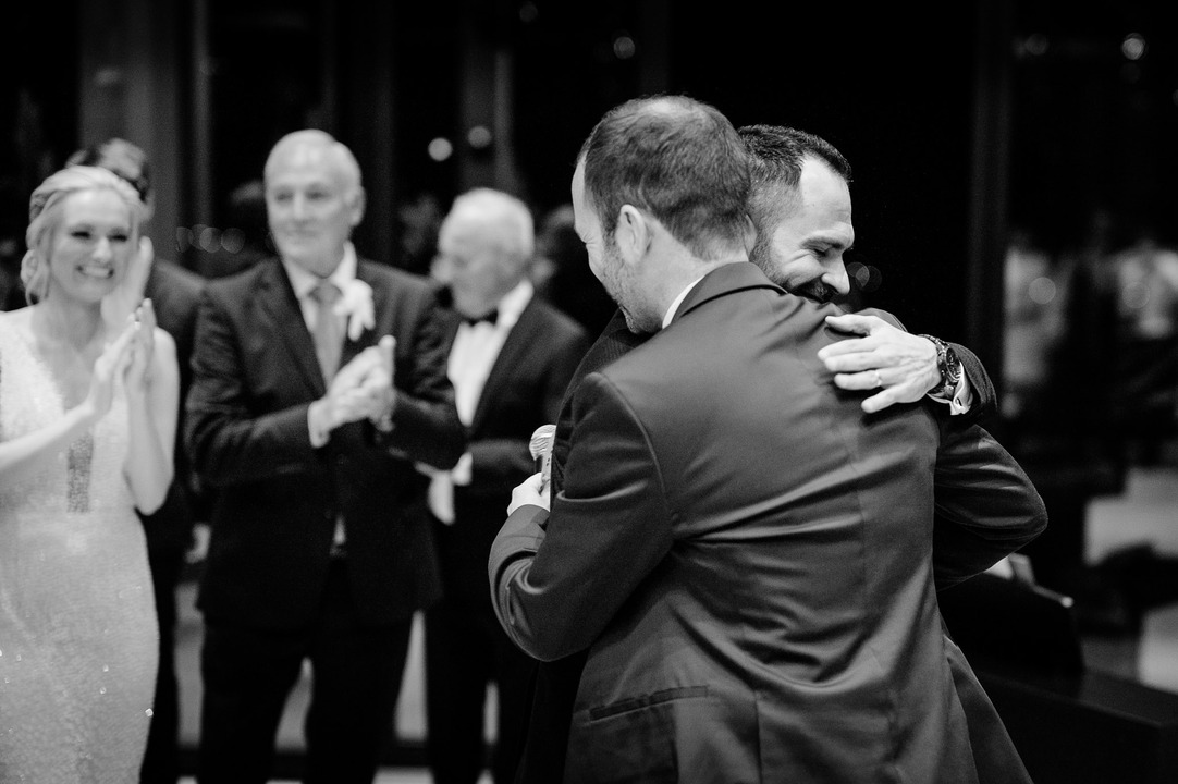 Groom hugging a guest while others applaud in the background.