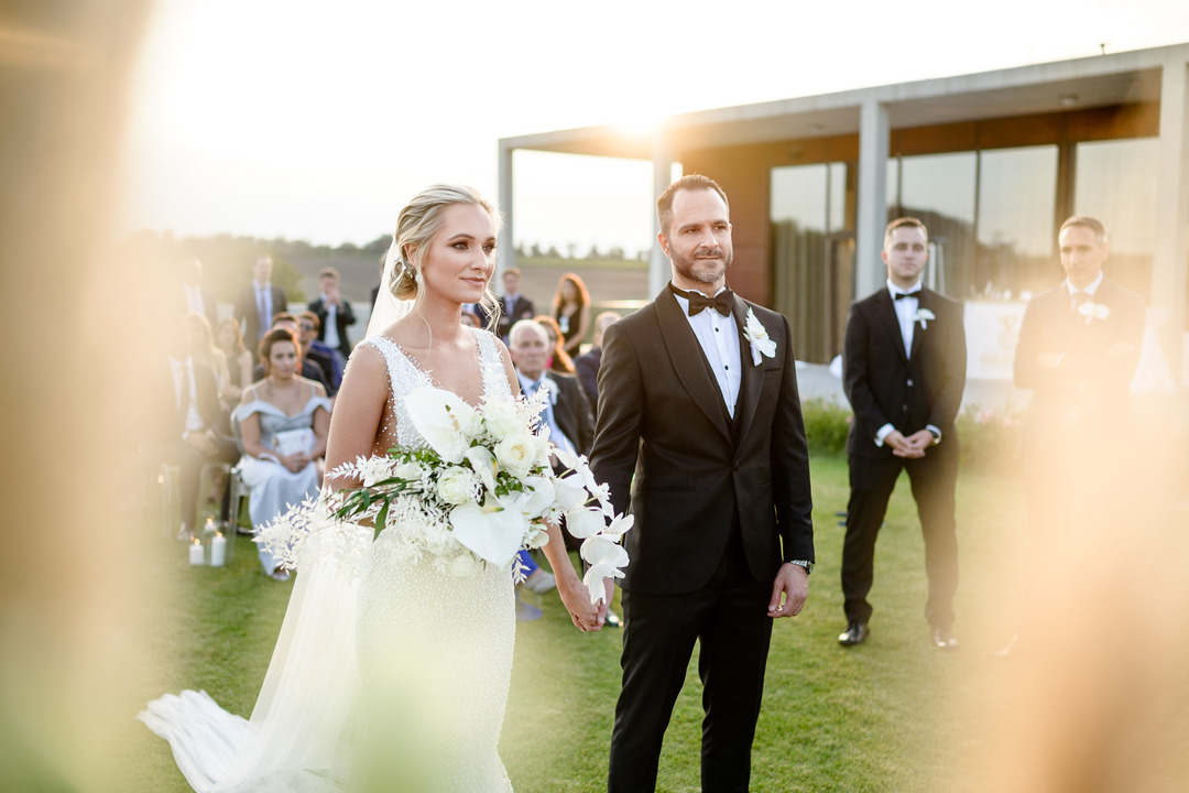 The groom and bride hold hands during the ceremony, with the evening sun peeking from behind the building.