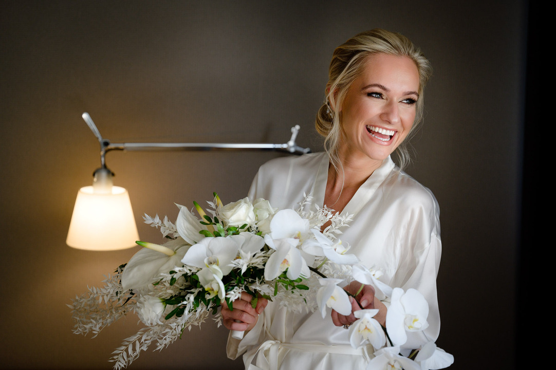 Bride in a white robe holding a bouquet of white flowers, smiling during the morning.