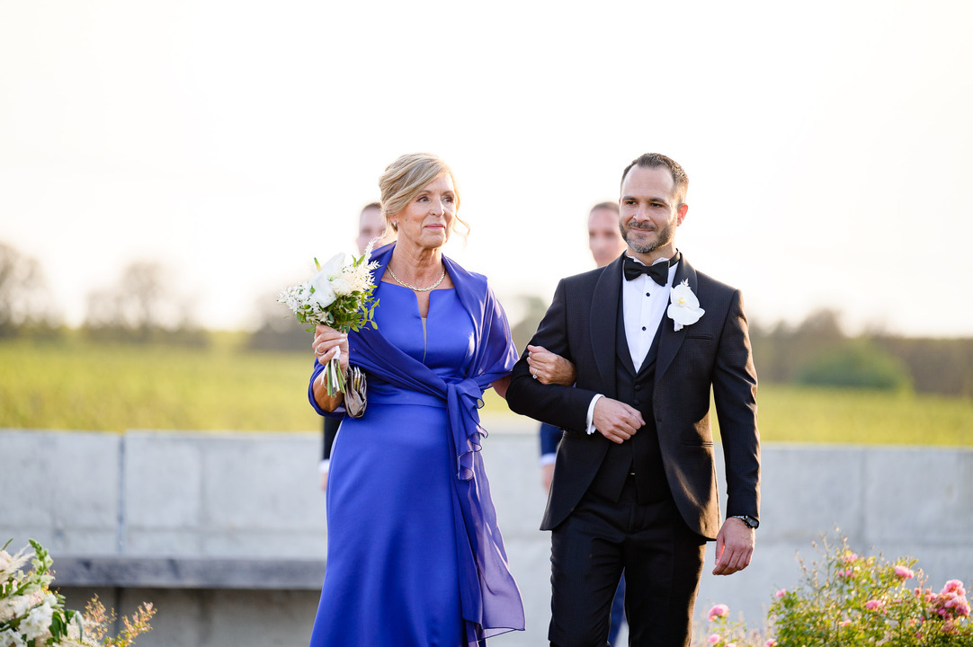 Groom walking with his mother, both dressed formally, at a winery wedding.