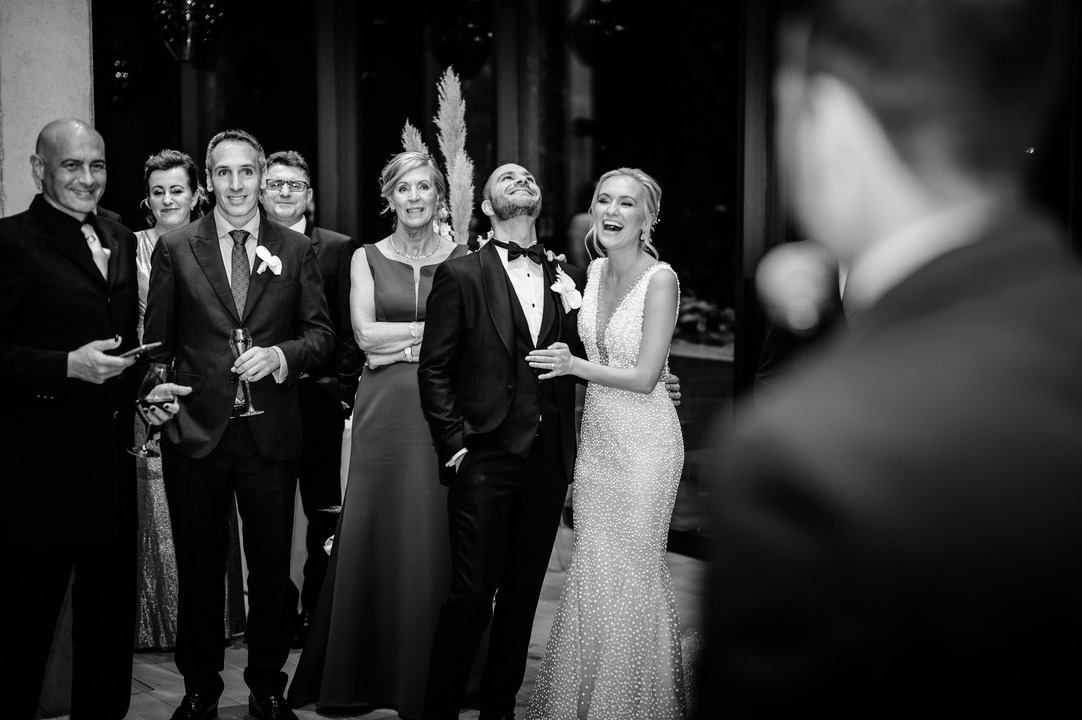 Best man giving a speech while guests and newlyweds smile.