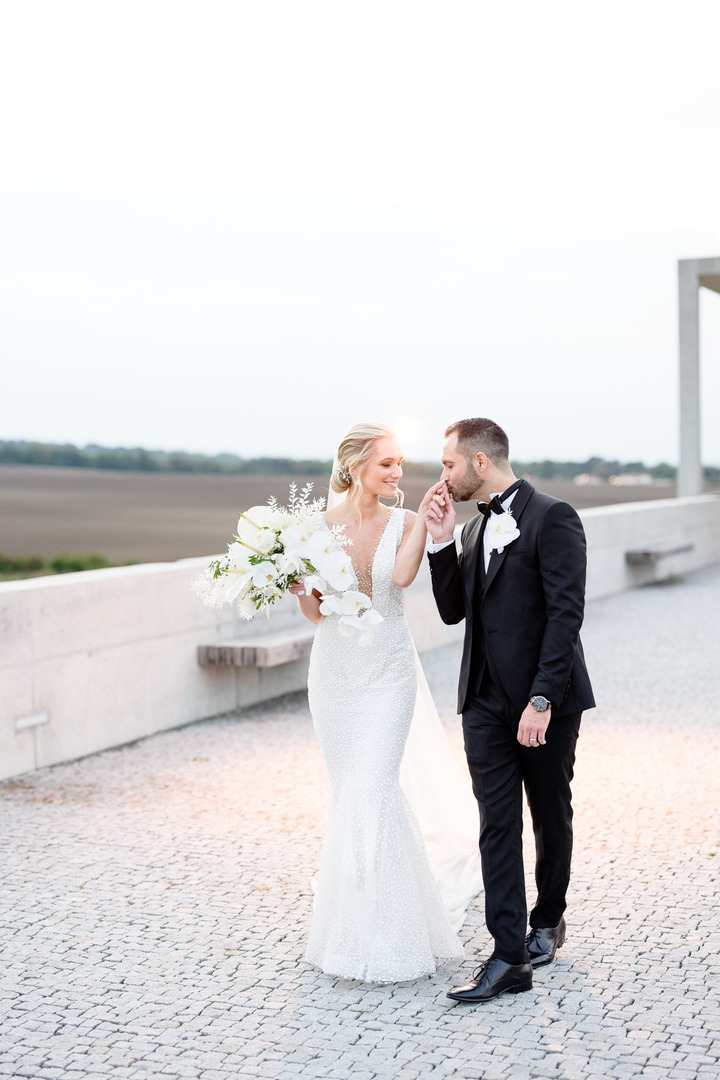  Groom tenderly kissing the bride's hand as they walk together at Obelisk Winery, with the bride holding a bouquet of white flowers. 