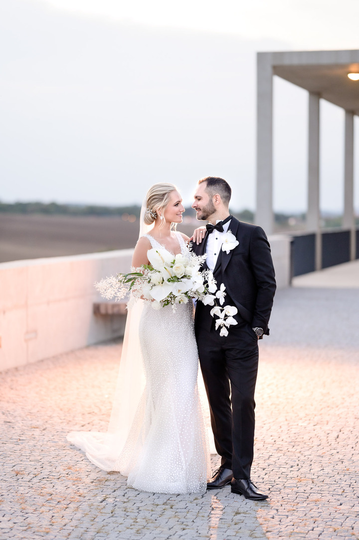 A bride and groom embrace, holding elegant white floral bouquets, with the bride in a lace gown and the groom in a black tuxedo.