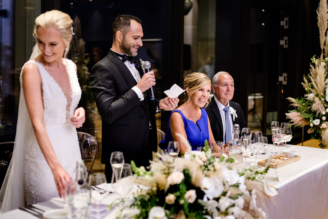 The groom speaks into the microphone while others laugh.