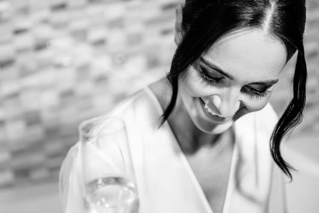 Black and white close-up of the bride’s face, holding a glass, her head slightly bowed and smiling happily.
