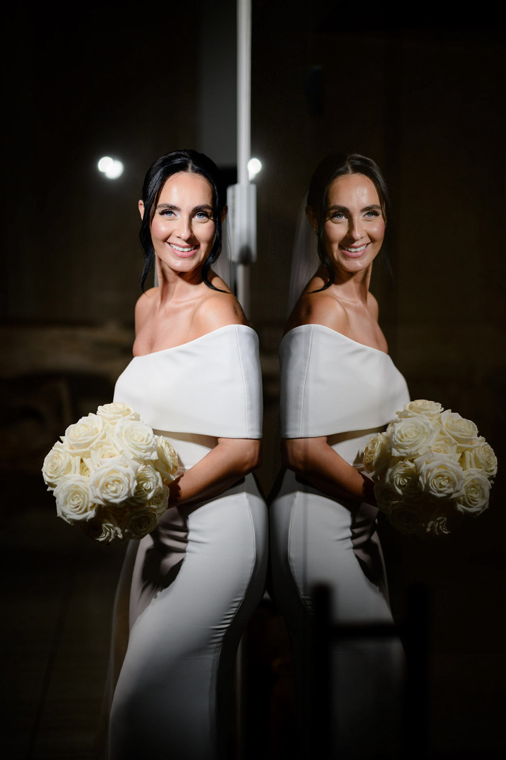 Beautiful bride in an elegant wedding dress with a bouquet of white roses inside Villa Cattaleya – a moment of pure joy and beauty, with her reflection visible in the glass she leans against.
