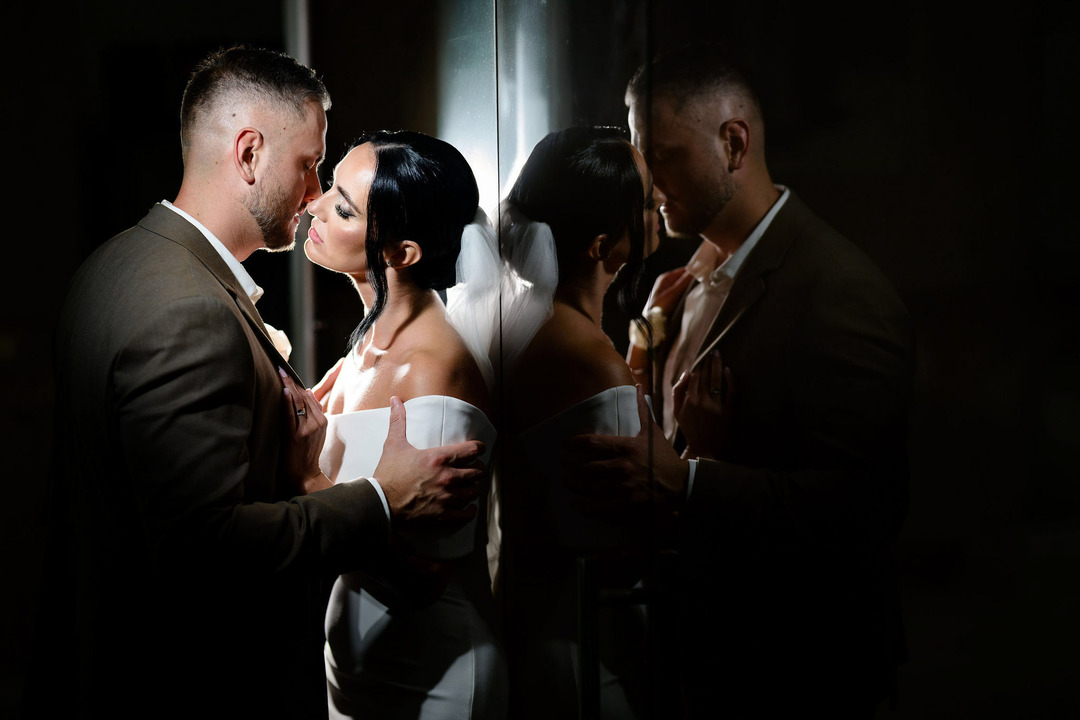 The groom and bride are in a passionate embrace, leaning against a glass wall, where their reflection is visible.
