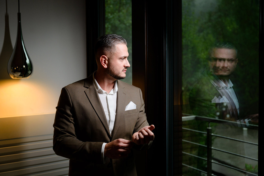 The groom, during his morning preparations, looks out the window, with his reflection visible in the glass.

