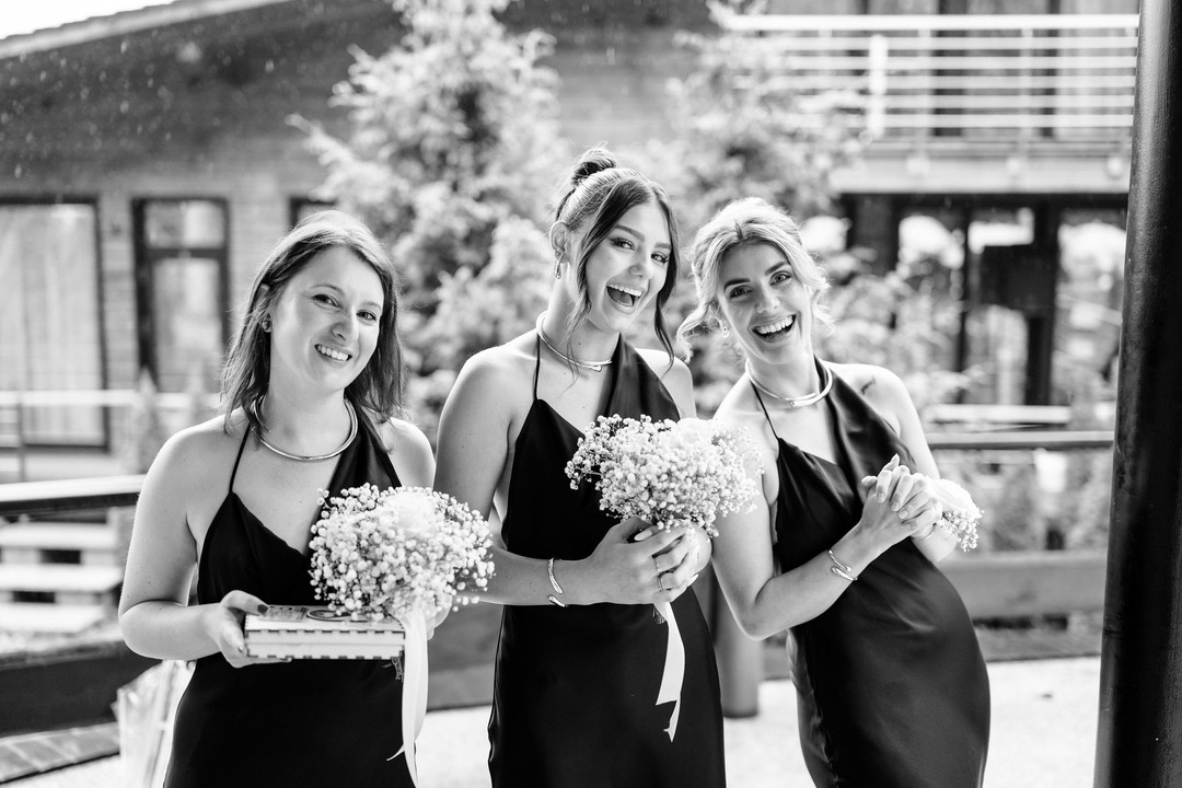 A black-and-white photograph of three of the bride's bridesmaids in black dresses, all laughing joyfully. They are holding bouquets in their hands.
