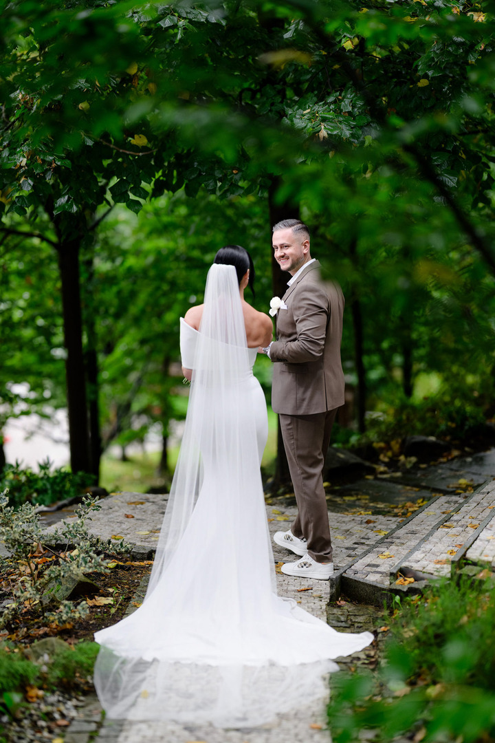 Photo showing the bride from behind, the groom turning toward her, reaching out his hand and smiling at her.